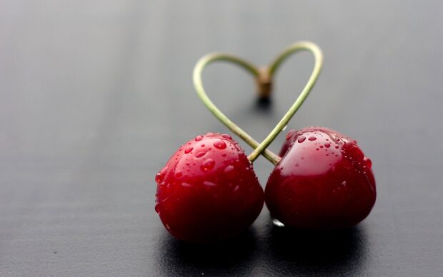 Two fresh cherries with stems forming a heart shape and water droplets on a dark surface