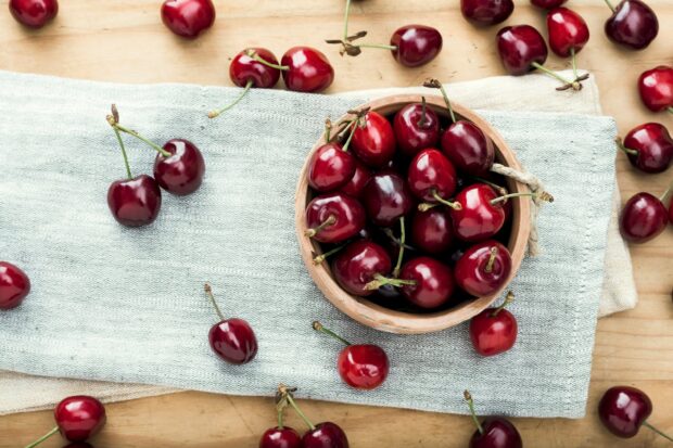 A bowl of fresh ripe cherry on a cloth with scattered cherries on wooden table