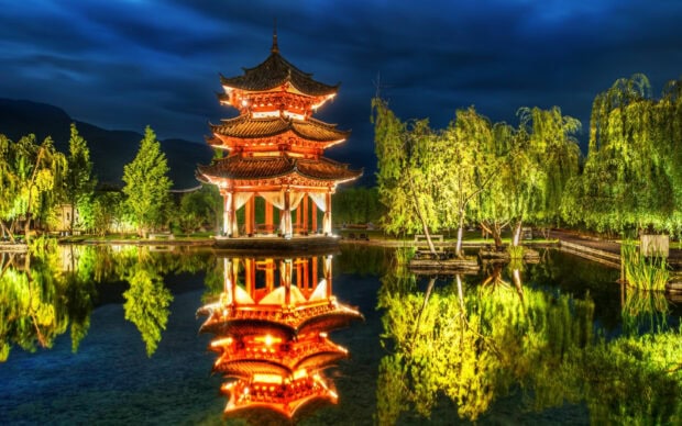 Traditional Chinese architecture illuminated at night reflecting in a calm water lake surrounded by lush green trees