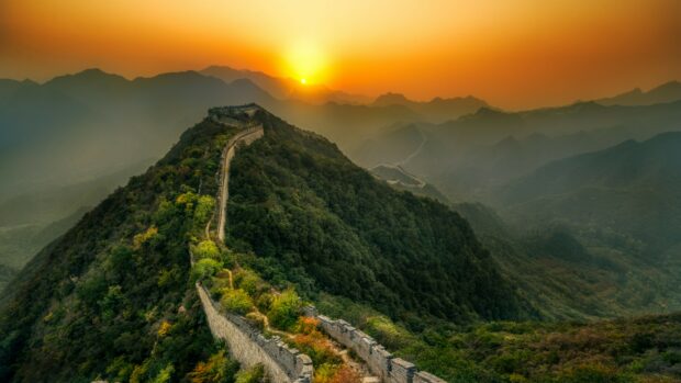 The Great Wall of China winding over lush green mountains at sunset with a vibrant orange sky