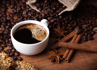 A close up of cinnamon sticks with coffee beans and a cup of black coffee on wooden surface