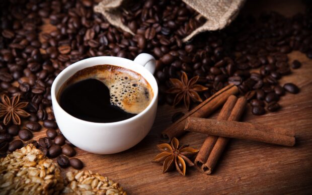 A close up of cinnamon sticks with coffee beans and a cup of black coffee on wooden surface