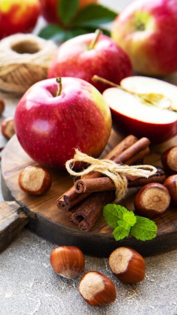 Fresh cinnamon sticks with red apples and hazelnuts on a wooden board surrounded by mint leaves