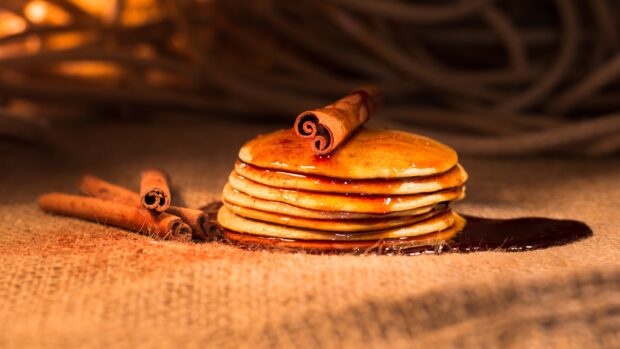 A stack of pancakes topped with a cinnamon stick and syrup on a burlap surface