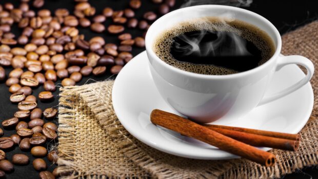 A hot cup of coffee with cinnamon sticks on a white saucer surrounded by coffee beans on burlap