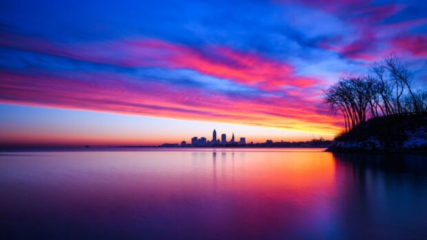Vibrant Cleveland skyline at sunset with colorful clouds over calm water and trees on the shore