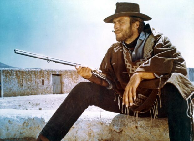 Clint Eastwood wearing a poncho and hat holding a rifle in a western desert scene