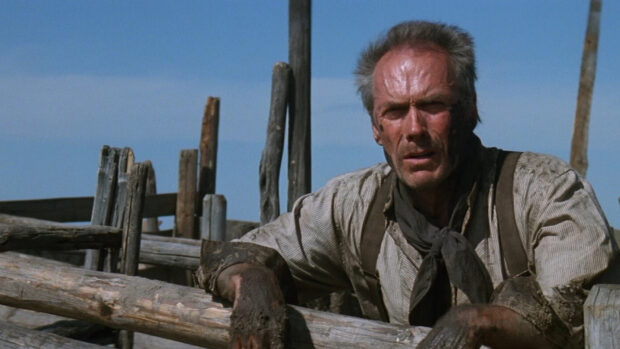 Clint Eastwood wearing work gloves leaning on wooden fence in rustic outdoor setting