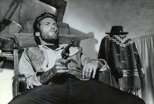 Clint Eastwood holding a revolver while reclining in a chair in a western scene