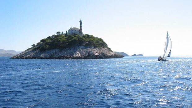 Island with lighthouse in Croatia surrounded by sea and a sailboat sailing nearby