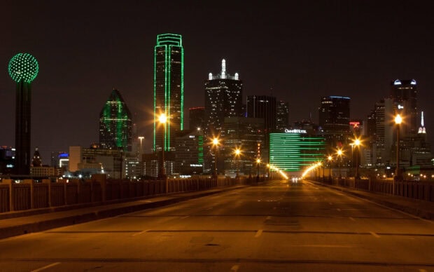 Night view of Dallas Texas city skyline with illuminated green buildings and empty road ahead