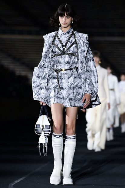 Female model wearing Dior patterned outfit with white boots and handbag on runway