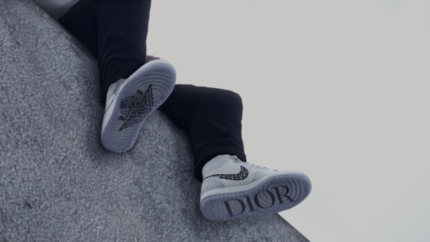 Person wearing Dior branded sneakers sitting on a gray surface with visible Dior logo on the soles