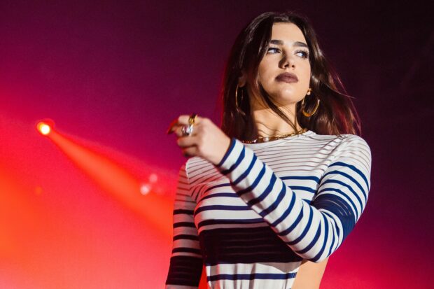 Dua Lipa wearing striped long sleeve top performing on stage with red and purple lights in the background