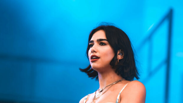 Dua Lipa performing on stage with blue background and wearing layered necklaces