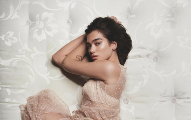 Dua Lipa lying on a patterned surface with a sheer beige dress and tattoo visible on her arm