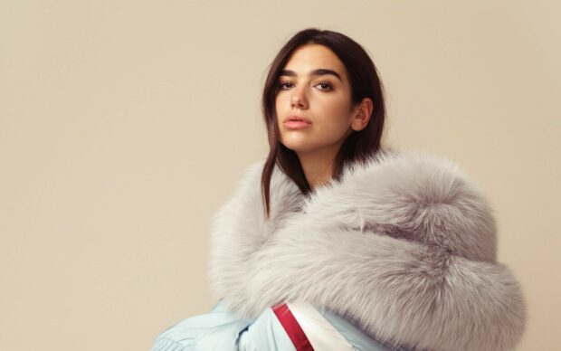 Dua Lipa wearing a light blue jacket with a large gray fur collar against a beige background