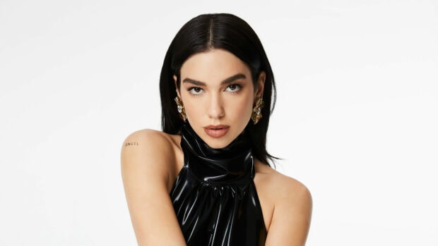Dua Lipa wearing a shiny black outfit with gold earrings and angel tattoo on shoulder