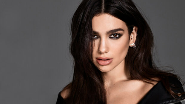 Dua Lipa with long dark hair and smoky eye makeup posing against a gray background