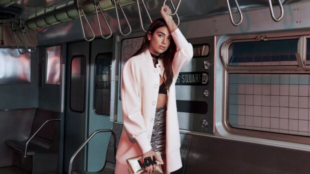 Stylish Dua Lipa posing inside a subway train wearing a white coat and metallic skirt
