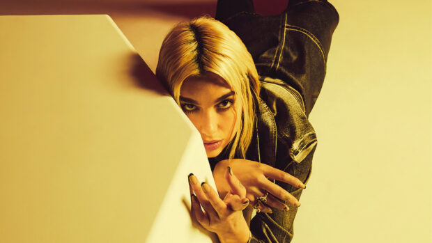 Dua Lipa posing with intense gaze and blonde hair leaning on a yellow surface in a stylish photo