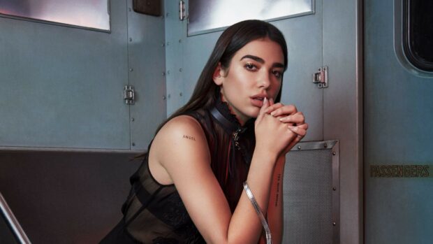 Dua Lipa sitting in a metallic room with her hands clasped wearing a sheer black top