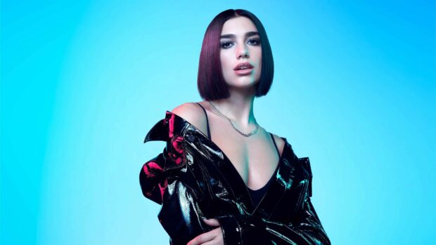 Dua Lipa wearing a black shiny jacket posing against a blue background