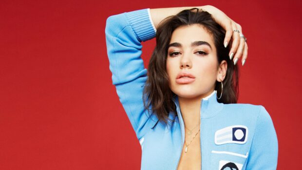 Dua Lipa wearing a blue jacket posing against a red background with a confident expression