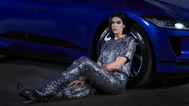 Dua Lipa wearing a sequined outfit sitting on the ground beside a blue car