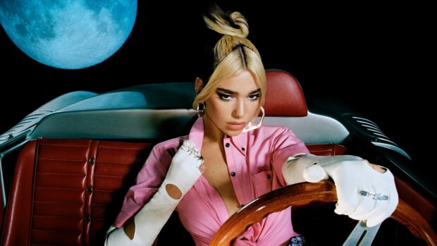 Dua Lipa wearing white gloves and a pink shirt sitting in a car with a blue moon behind her