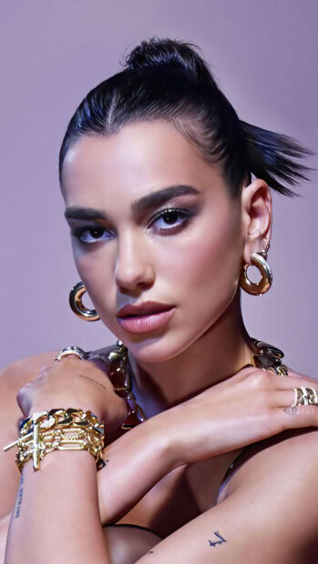 Dua Lipa wearing gold jewelry and makeup with her hair tied back in a close up portrait