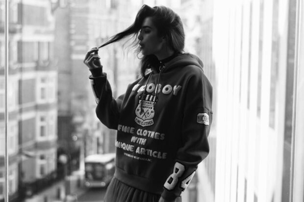 Dua Lipa standing by the window playing with her hair in a casual hoodie