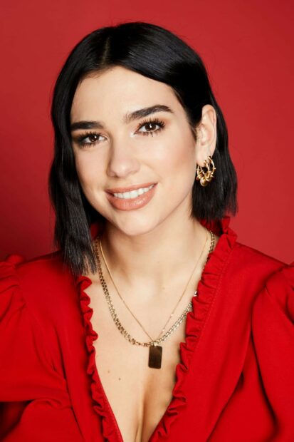 Dua Lipa smiling with short black hair wearing red outfit and gold jewelry on red background