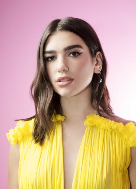 Dua Lipa wearing a yellow dress with natural makeup on a pink background
