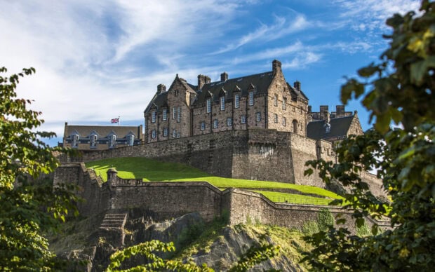Historic Edinburgh Castle standing atop a green hill surrounded by trees under a blue sky