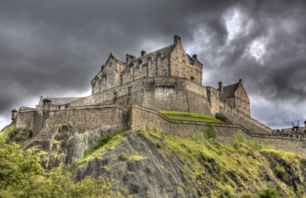 Edinburgh Castle stands majestically on the rocky hill surrounded by green vegetation and cloudy sky