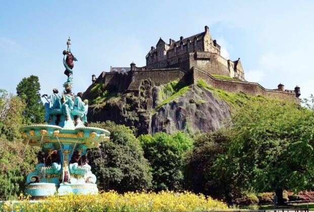 Edinburgh Castle stands prominently above lush greenery and a decorative fountain in a vibrant garden setting