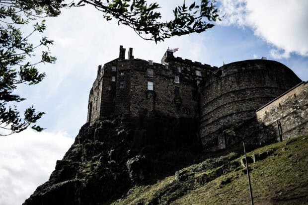Edinburgh Castle stands prominently on a rocky hilltop surrounded by greenery and a bright sky