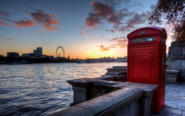 A red telephone booth beside the River Thames in London at sunset with the London Eye in the background