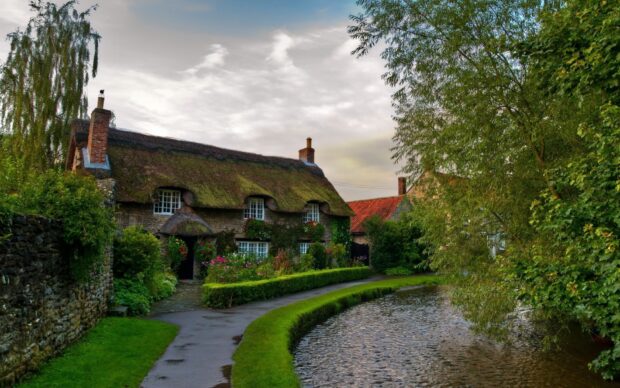 A charming England cottage with a mossy roof beside a winding river and lush greenery