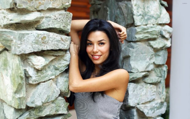 Eva Longoria smiling and posing by a stone wall with her arms raised above her head