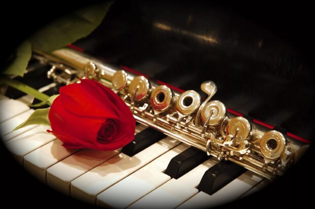 A flute resting on piano keys with a red rose placed on top of the keys