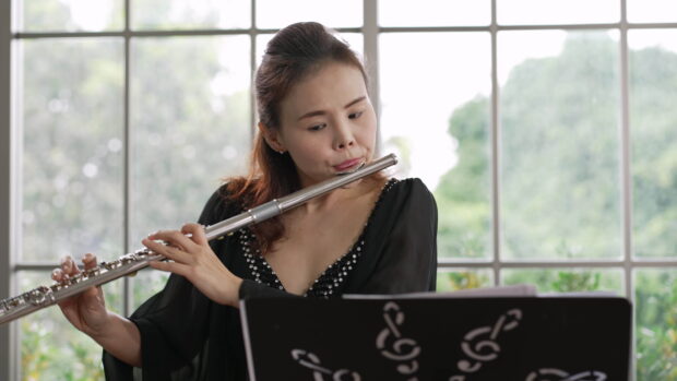 Woman playing flute with focused expression indoors near window and music stand