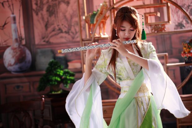 A woman wearing traditional clothing playing a decorative flute indoors with an artistic background