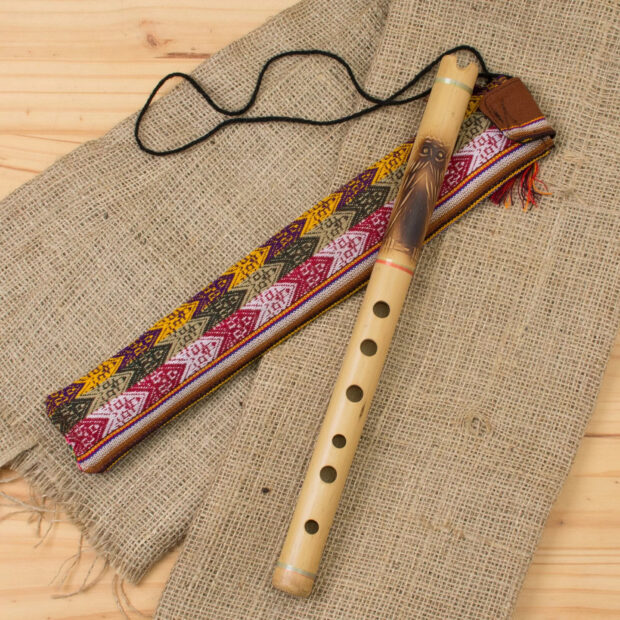 A wooden flute with carved owl design lying on a textured fabric with a colorful patterned strap