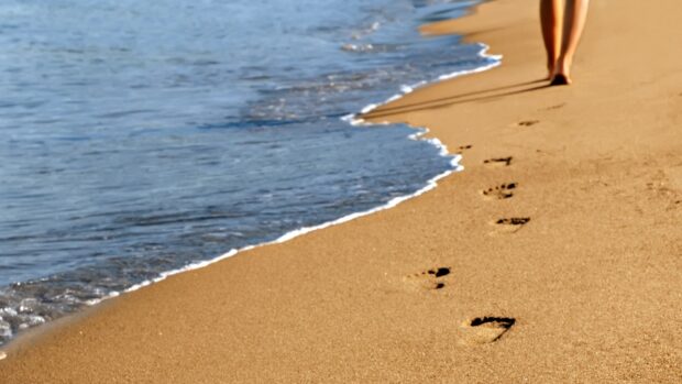 Footprints in the sand along the shore near the ocean with a person walking nearby