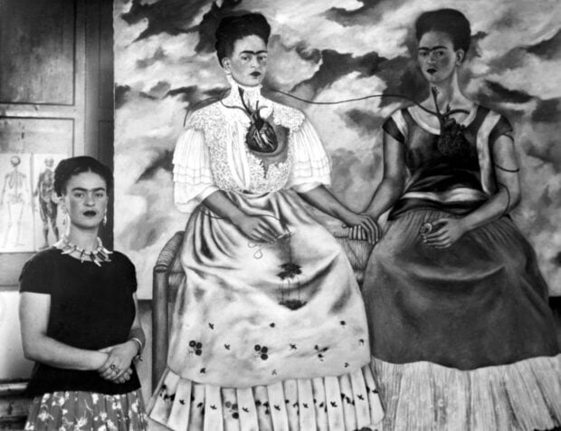 Frida Kahlo standing next to her surrealist painting featuring two women connected by a heart
