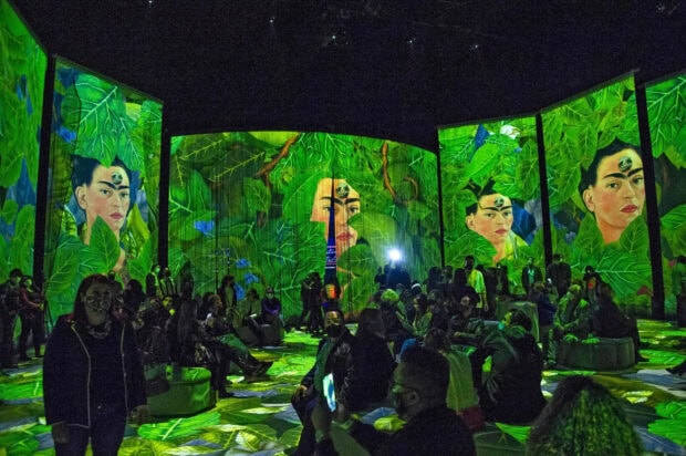 Frida Kahlo surrounded by lush green leaves in an immersive art exhibition with many visitors seated