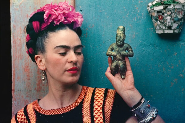 Frida Kahlo holding a small ancient statue with traditional hair and bright flowers