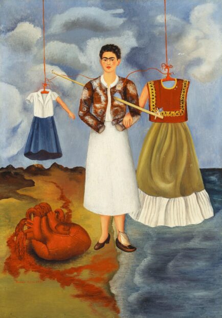 Frida Kahlo standing between two dresses with a large heart on the ground and a cloudy sky background
